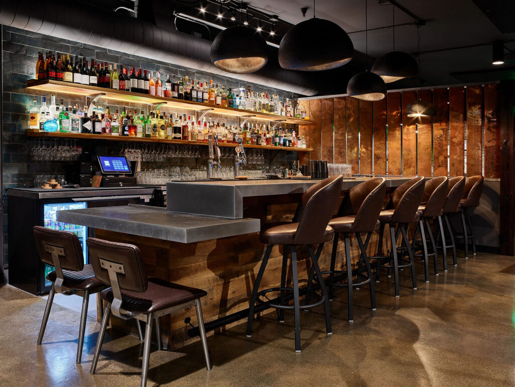 A bar with two shelves full of alcohol bottles. Six leather swivel barstools and two brown leather seats sit empty at the bar which is topped with zinc counters. Copper panels line the far end and five pendants of varying sizes hang from the ceiling.