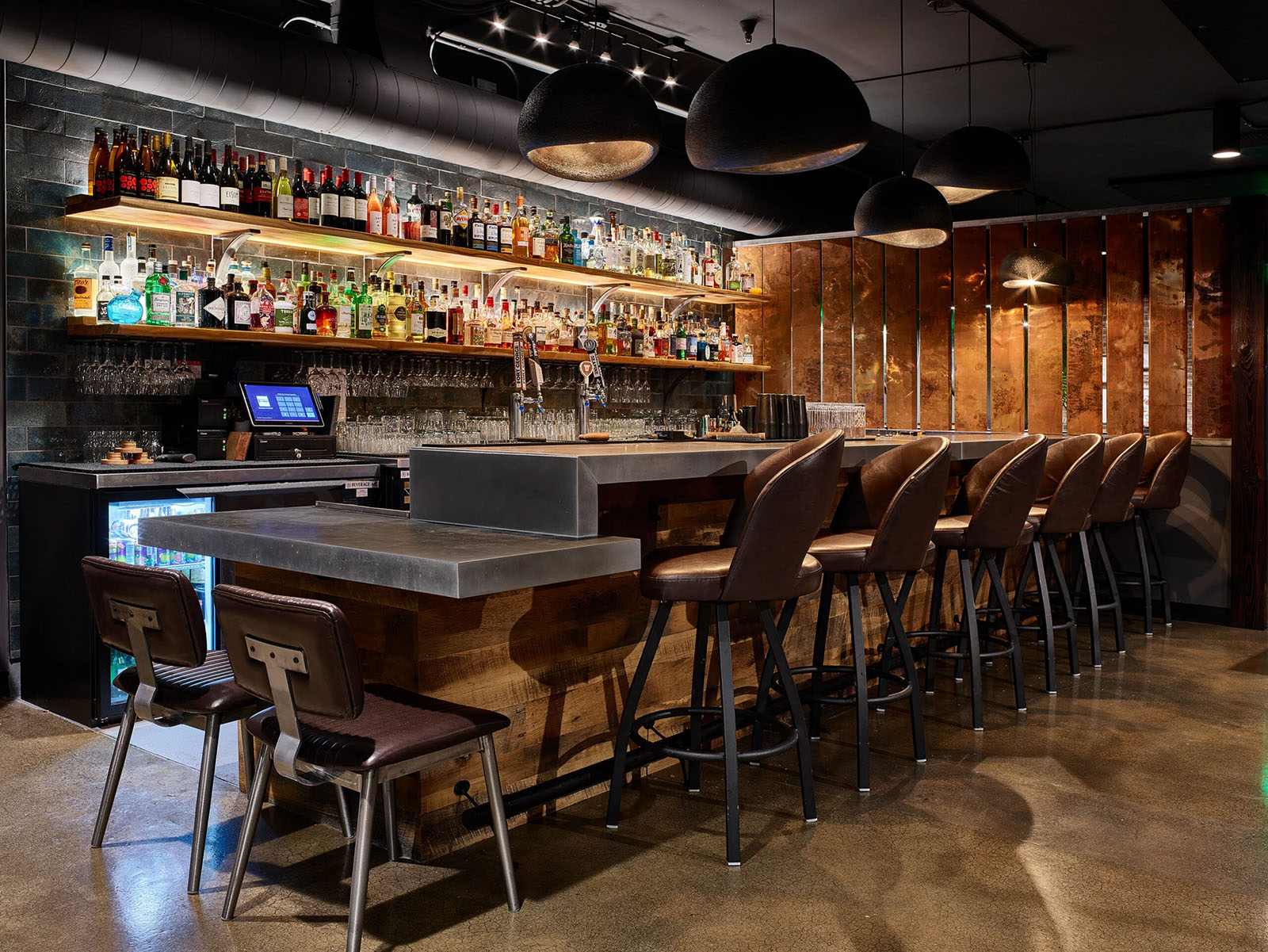 A bar with two shelves full of alcohol bottles. Six leather swivel barstools and two brown leather seats sit empty at the bar which is topped with zinc counters. Copper panels line the far end and five pendants of varying sizes hang from the ceiling.