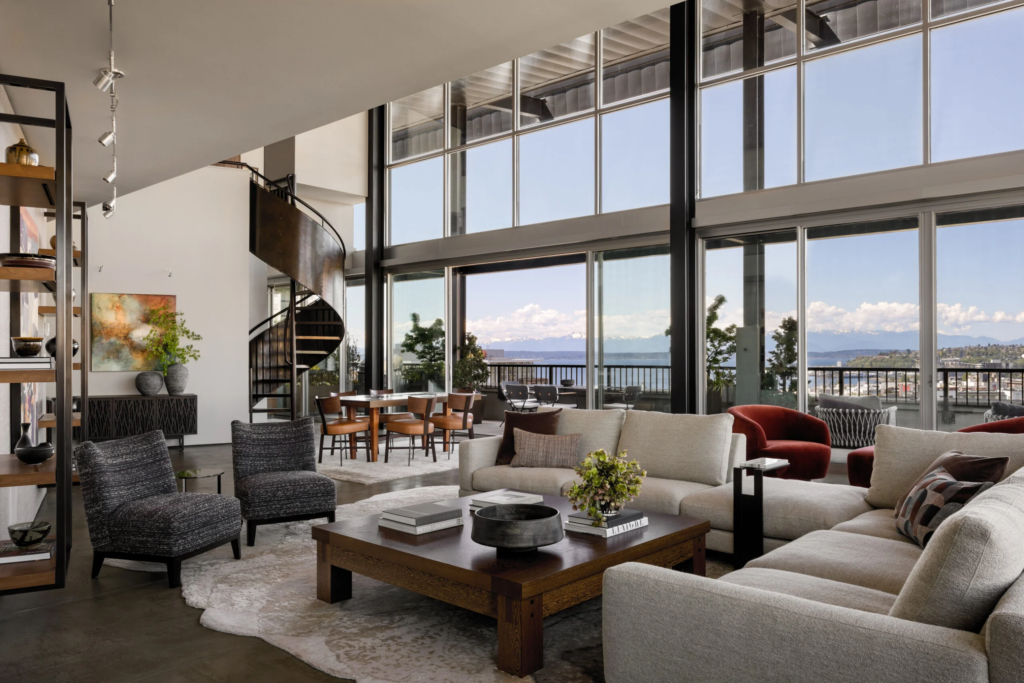 A modern penthouse apartment with floor to ceiling windows overlooking Elliot Bay in Seattle. A large cream colored sectional sofa sits across from two dark grey armless chairs. A low, dark wood coffee table holds books, a dark grey bowl, and a green plant. A bookshelft on the left, two red swivel chairs behind the sofa on the right. A wood dining table with 6 chairs with tan leather seats sits adjacent to a custom metal spiral staircase ascending to an upper floor.