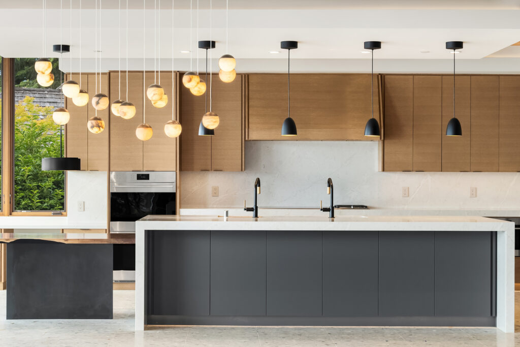 A kitchen with a large island features white marble waterfall counters, a mix of mid-tone wood and dark cabinets. Four black pendant lights hang above the island. A chandelier eighteen gold colored glass globes hangs in the foreground.