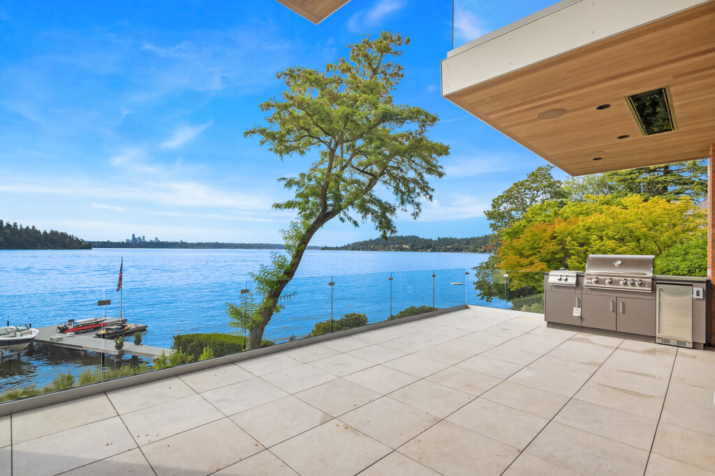 A patio with large stone pavers, stainless steel grill, and glass railings overlooking the lake and Seattle skyline in the distance