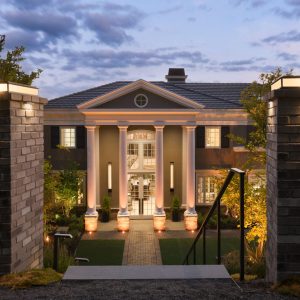 Two brick posts flank the approach to a Georgian style mansion with four white columns, brown stucco and grassy front yard. A modern geometric chair and ottoman sit atop a green grass yard. It is sunset and light shine up the columns.