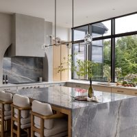 A grand kitchen with grey marble counters and backsplash, plaster hood over the cooktop, light wood and white custom cabinets, and a wall of windows behind the sink look out to a greenery and purple flowers. The counters have a variety of fruits, cookbooks, a bottle of champagne and two pink coupe glasses.
