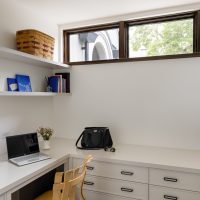 A small office nook with built-in white cabinetry. A laptop, purse, sunglasses, and small bouquet of pink flowers in a white base sits atop the counter. Shelves on the wall hold books and a wicker basket.