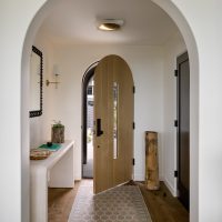 An interior view of an entry vestibule to a home looking towards the front door. The door and entry are arched. The floor is medium brown hardwood with wide planks. A long white console table sits to the left, holding a plant, book, and marble bowl. Above the table is a mirror and a brass wall mounted sconce with a white lampshade