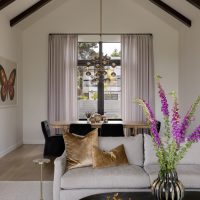 An interior home view features an oval shaped coffee table holding a large vase with vibrant purple flowers, a book with three gold bunny statues sit atop. A beige sofa with two gold velvet pillows sits mid image with a small drink table to it's left. In the background is a light wood dining table with six black upholstered armless chairs. The walls are white and the high pitched ceiling feature dark wood beams