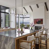 A grey marble kitchen island holds a wine bottle, two glasses, and a tray. Three wood chairs sit at the island with beige grey cushioned sets. The view in the background through the adjacent living room peeks at Elliot Bay and the Seattle skyline.