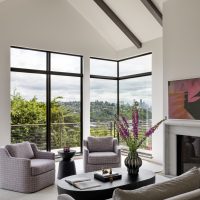 A living room with large floor to ceiling windows overlooks Elliot Bay and the Seattle skyline. The walls are white and contrasting dark wood beams line the high pitched ceiling.