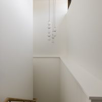 A view of a residential staircase looking down from the top. Walls are white, a wood hand rail with black metal brackets on the left and a chandelier above. The chandelier is antique brass circle at the ceiling, with three strands of white bulbs hanging in varying lenghts.