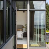 A view from a wood patio looking into an open glass door of a bedroom. The bedroom has floor to ceiling windows.