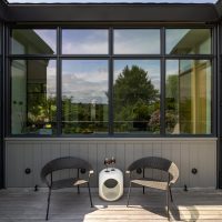 A wood patio holds two woven chairs, a small white table holding two wine glasses. Black metal windows and doors surround the patio.