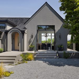 A modern Tudor style home with grey wood vertical siding, arched front door entry, and black metal windows. Two french doors are open to the dining room inside.