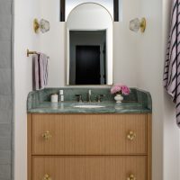A wood bathroom vanity with a green marble top holds a bouquet of pink flowers. An brass framed arched mirror is flanked by two brass and glass wall sconces. Pink towels with navy stripes hang on the left and right sides.