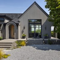 A modern Tudor style home with grey wood vertical siding, arched front door entry, and black metal windows. The front yard is xeriscaped with pebbles, ornamental grasses and a large tree fills the right side.