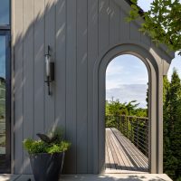 An arched entry to a wood patio on a modern Tudor home. Grey vertical wood siding clads the house and a modern black metal and glass light fixture hangs over a large decorative planter filled with flowers and plants.