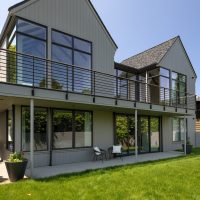 A two story house with grey vertical cedar plank siding, black windows, two pitched gable roofs, black metal railings on the deck and grassy yard