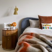 A soft wool blanket in orange, brown, cream and blue plaid is draped across a bed with a cushioned navy blue headboard. A wood cabinet in an pentagonal shape hold a white picture frame and a bouquet of orange Gerber daisies sits to the left of the bed. A brass wall sconce hangs on wall above table.