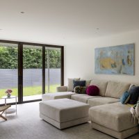 A beige sectional sofa holds a variety of colorful throw pillows in blues, green, and magenta. A green wood checked throw blanket drapes over the arm on the ride side of the sofa. An armless woven wicker and wood chair sits across from the sofa aside a white metal round table with a pink bouquet on top. A wall of sliding window doors look out to a green grass yard with a grey fence. A large abstract print hang on the wall above the sofa in colors of blue, yellow, and green.