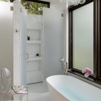A luxurious bathroom features a free standing modern white tub, a walk in shower with frosted glass door, white square tiles, built in shelves hold a few bottles of toiletries. A bubble shaped chandelier hangs over the tub. A large window to the right of the tub holds a small bouquet of pink flowers. A clear acrylic chair sits to the left with a pink flower towel draped across the seat.