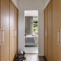 A corridor view looking into a bedroom. Four wood closet doors line the sides with long clear acrylic pulls accented with brass. Wide plank wood floors hold a basket holding a blanket and a pair of white single strap sandles.