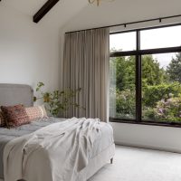 A peaceful bedroom view. Soft blankets in neutral beige and grey are draped across a bed with a few colorful printed pillows with orange and brown tones. A large brass chandelier hangs from a vaulted ceiling with dark wood beams. The black metal windows look out to lush green landscaping. Beige silk curtains hang on the left side of the window.