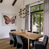 A dining room with a light wood table surrounded by six black velvet upholstered chairs. A large art print of a butterfly is on the white wall. Grey silk curtains flank a pair of glass french doors. An ornate brass chandelier with white glass globes hangs over the table from the vaulted ceiling with dark wood beams.