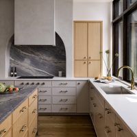A grand kitchen with grey marble counters and backsplash, plaster hood over the cooktop, light wood and white custom cabinets, and a wall of windows behind the sink look out to a greenery and purple flowers. The counters have a variety of fruits, cookbooks, and a flower arrangement