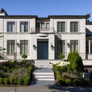 A large home with cream colored stucco, brown windows and roof, and a blue double front door. Flowers and bushes line both sides of the approach.